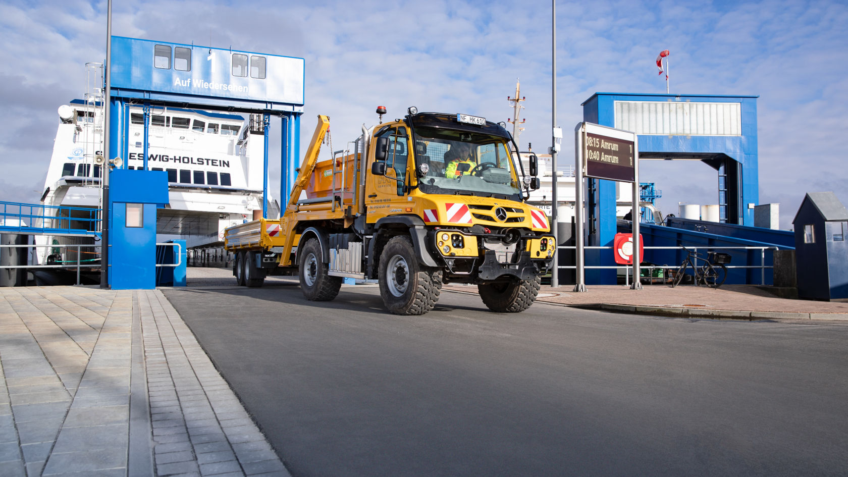 A yellow Unimog with a trailer driving off a ferry. The ferry, bearing “SCHLESWIG-HOLSTEIN” lettering, is moored at a blue ferry dock. The area in front of the dock is asphalted, and there is a paved walkway next to it.