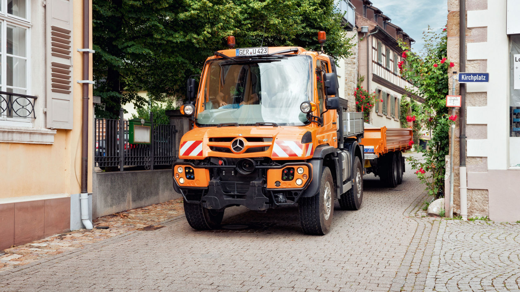 Ein orangefarbener Unimog mit Anhänger fährt auf einer gepflasterten Straße in einer Stadt.