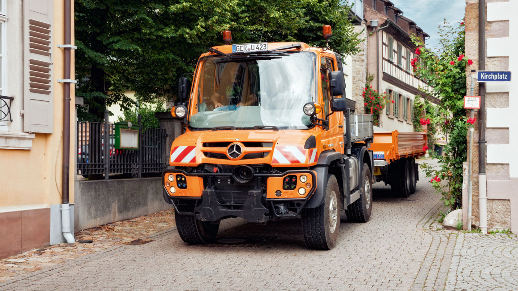 An orange Unimog truck with a trailer driving on a cobbled road in a village. A road sign reading “Kirchplatz” is attached to a building. Red roses trail over a wall next to the building. On the left side of the picture is a part of a house with shutters.