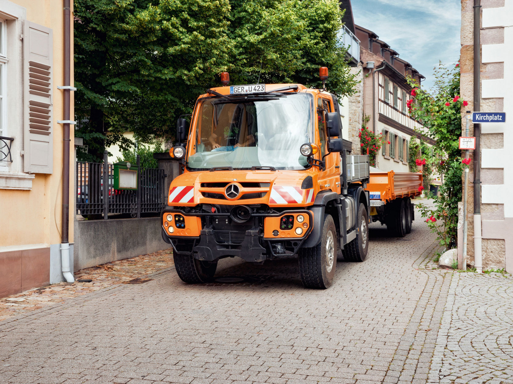 Ein orangefarbener Unimog Truck mit Anhänger fährt auf einer gepflasterten Straße in einer Ortschaft. An einem Gebäude ist ein Straßenschild mit der Aufschrift „Kirchplatz“ angebracht. Neben dem Gebäude ranken rote Rosen an einer Mauer. Links im Bild ist ein Teil eines Hauses mit Fensterläden zu sehen.