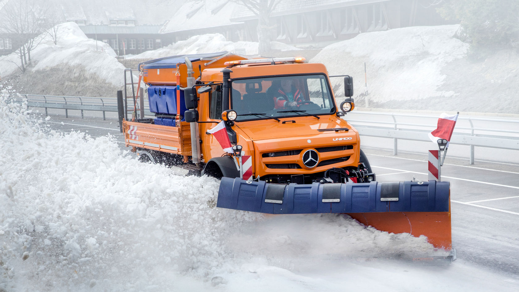 An orange Unimog with a snow plough clearing a snow-covered road. The snow is spraying to the side as the Unimog is driving through the white landscape. Snow-covered areas and a building can be seen in the background. The truck has a blue gritting salt container on its loading surface, and red and white warning bars on the snow plough.