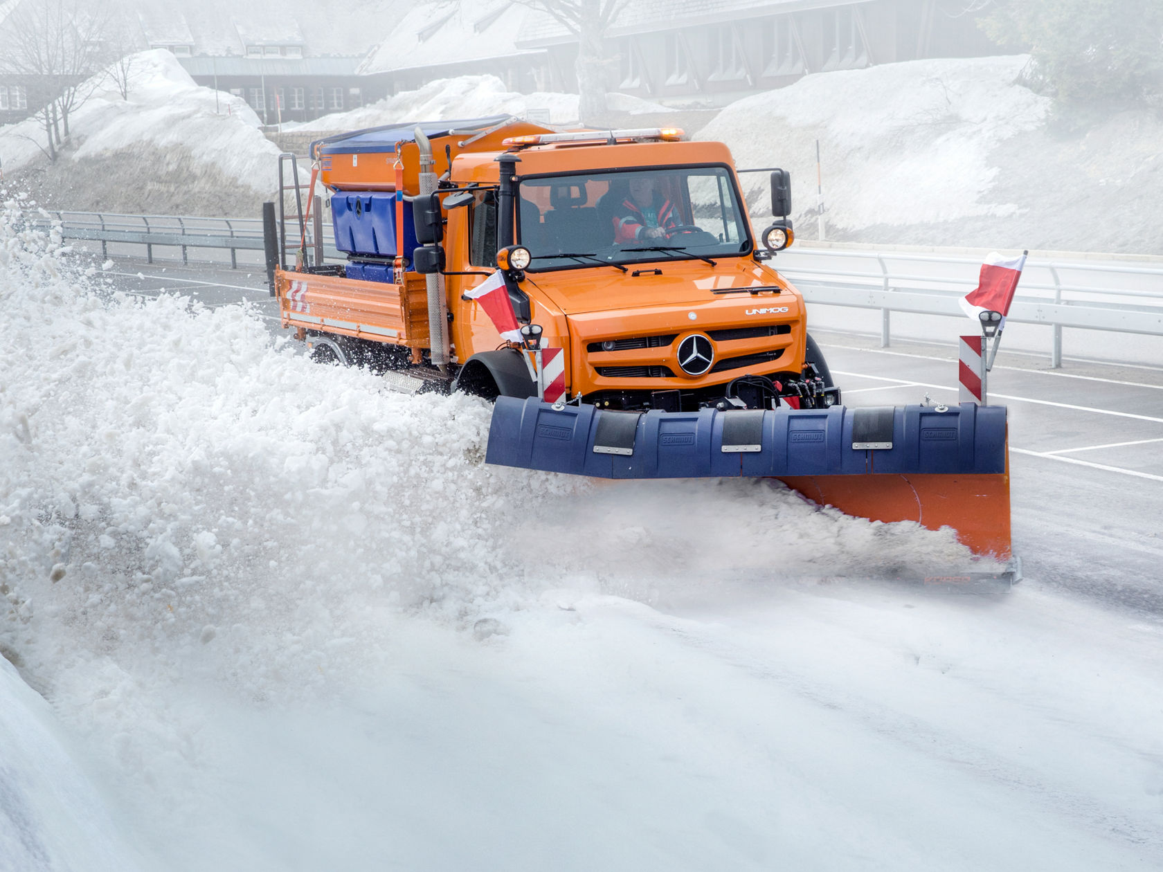 Ein orangefarbener Unimog mit Schneepflug räumt eine verschneite Straße. Der Schnee spritzt zur Seite, während der Unimog durch die weiße Landschaft fährt. Im Hintergrund sind schneebedeckte Flächen und ein Gebäude zu erkennen. Der Truck hat einen blauen Streusalzbehälter auf der Ladefläche und rot-weiße Warnbalken am Schneepflug.