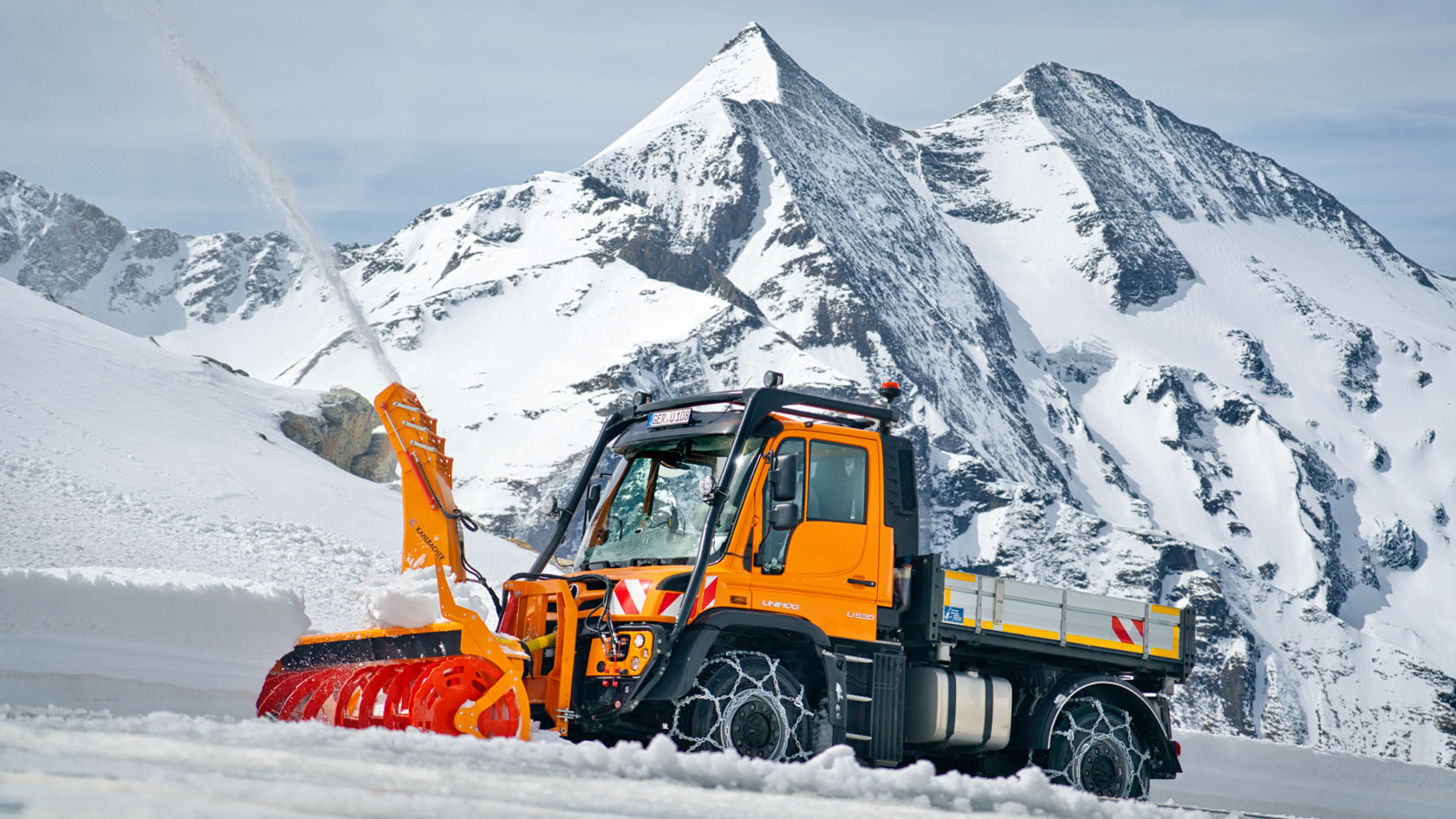 The image shows an orange Unimog with a snow blower clearing a snow-covered track in the mountains. The snow blower throws the snow high into the air. Snow chains are fitted on the tyres. Snow-covered mountain peaks are visible in the background.