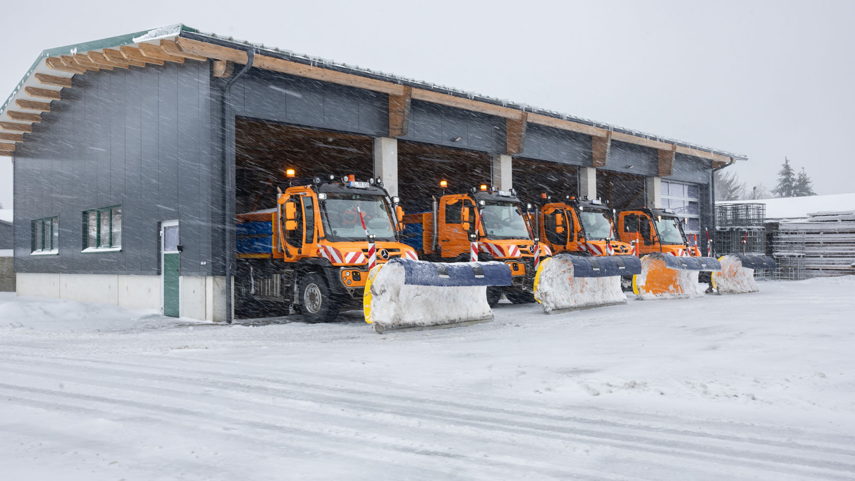 Three orange-coloured snow ploughs from Mercedes-Benz are ready to drive away in a vehicle hall. The snow ploughs are equipped with dozer blades and the surrounding area is covered in snow. It is snowing heavily.