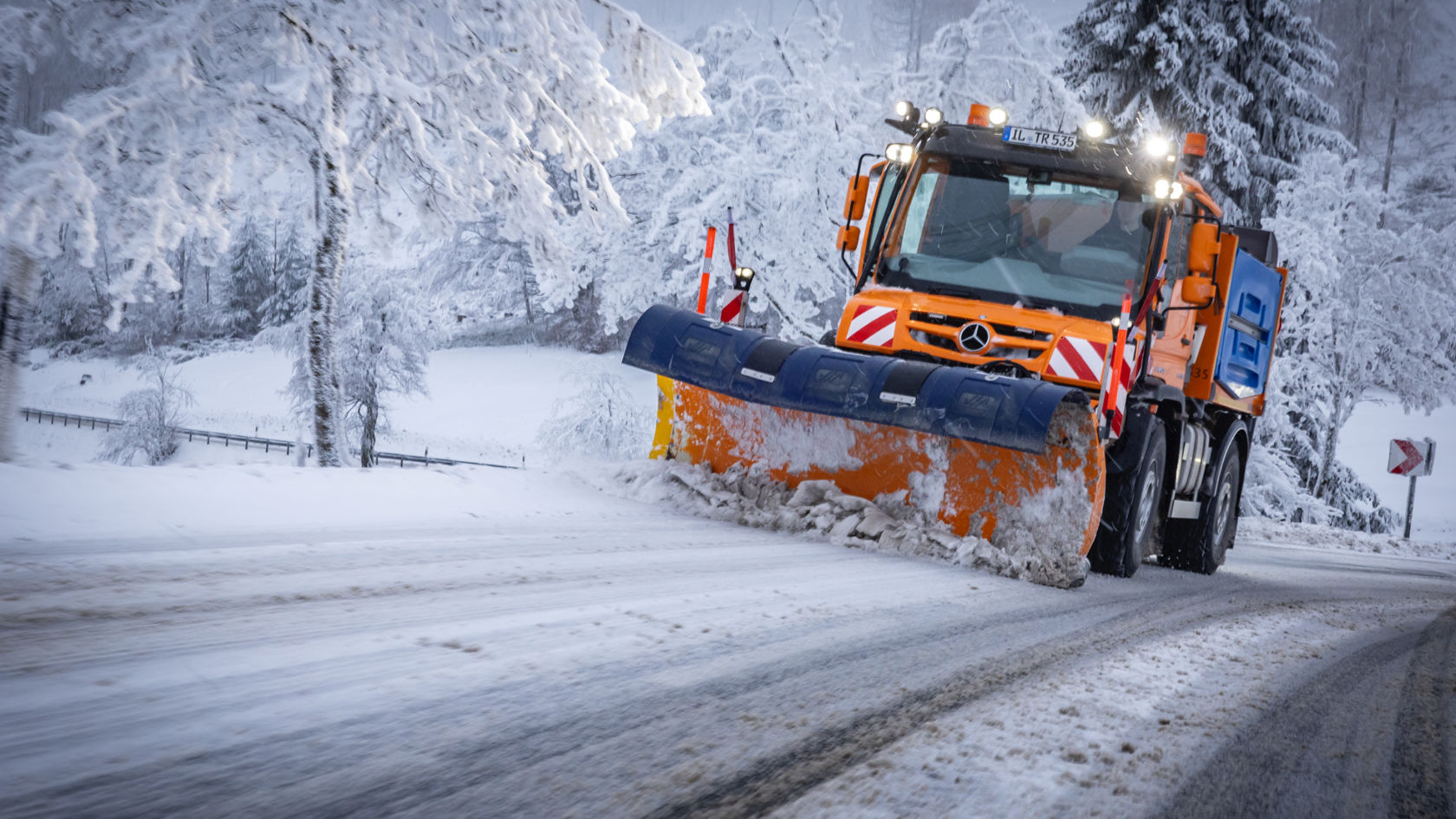 An orange Unimog snowplough driving on a snow-covered road clearing snow with its blue-orange snow blade. The road is covered with a mixture of snow and mud and has lane markings. Snow-covered trees line the edge of the road. 