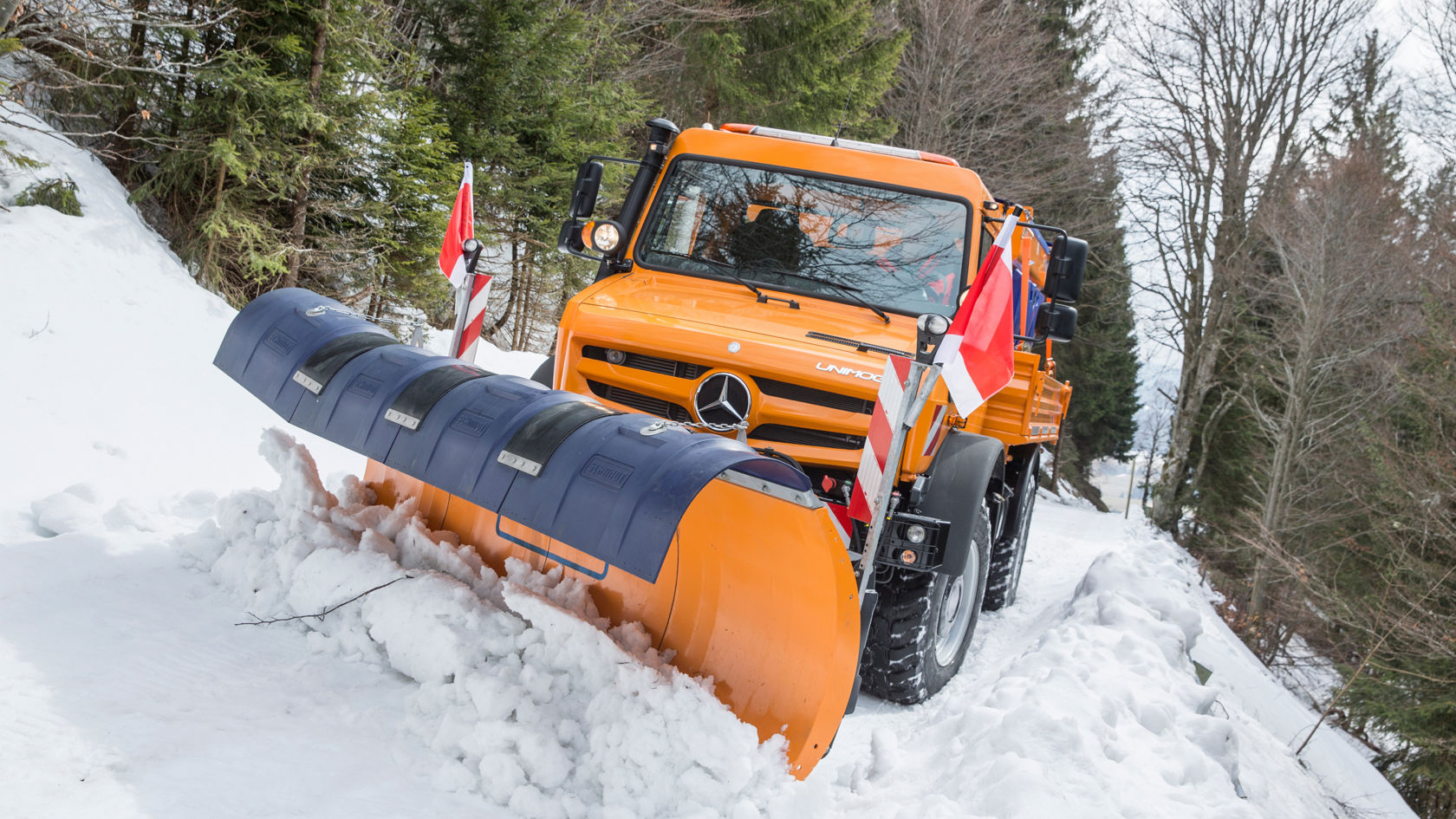 An orange Unimog with a snow plough attached clearing a path in a snowy landscape. Red and white warning flags are attached to the sides of the Unimog. Snow-covered trees and a wooded slope can be seen in the background. The sky is grey and the Unimog is not covered in snow. 