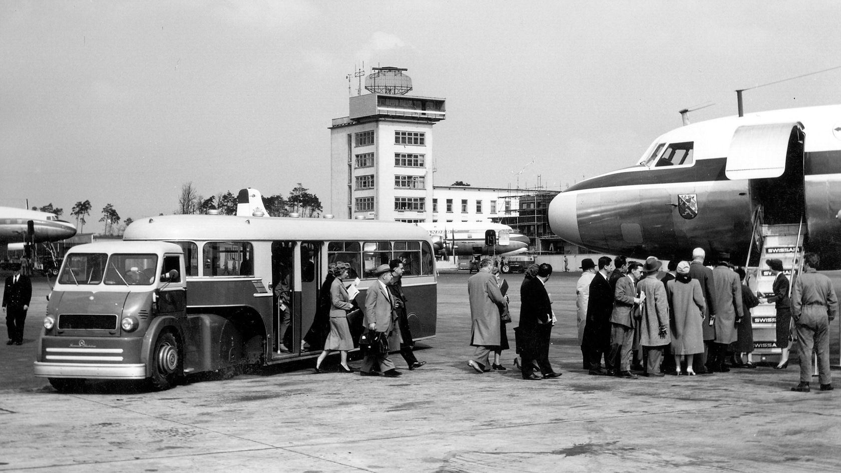 Flughafen-Szene mit Passagieren, die aus einem Bus aussteigen und in ein Flugzeug einsteigen, im Hintergrund ein Kontrollturm und verschiedene Flugzeuge.