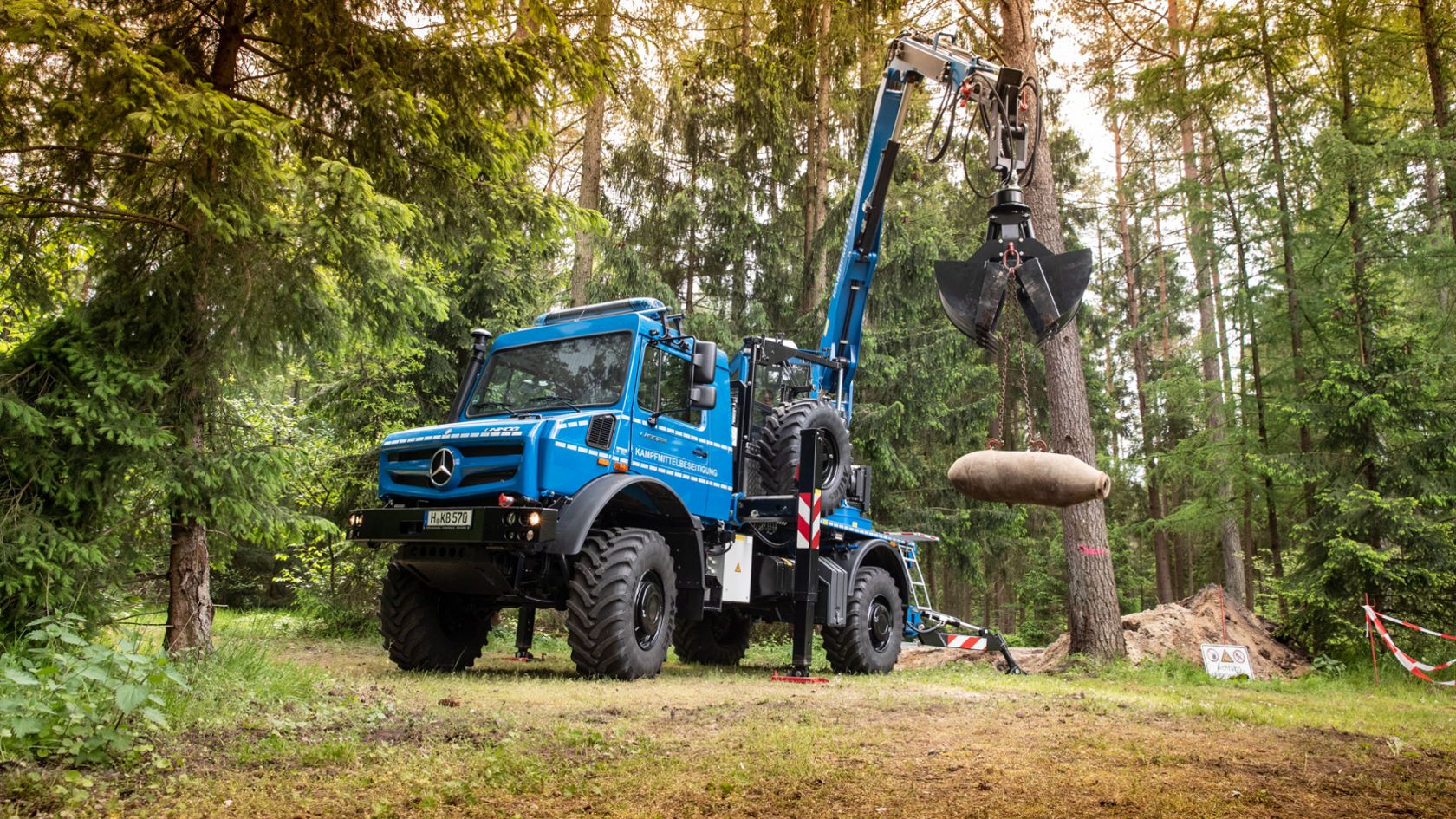 Un Unimog bleu avec bras de grue soulève une grande charge cylindrique dans une forêt.