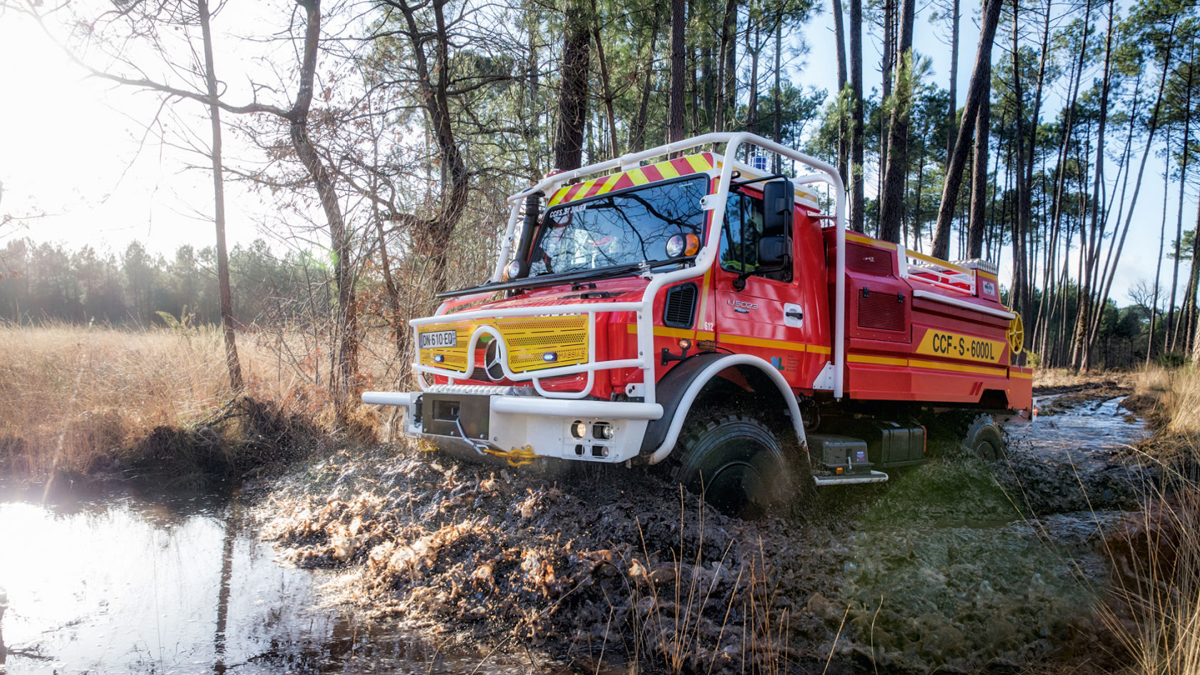 Červenožluté hasičské vozidlo Unimog projíždí bahnitou vodou v lese.