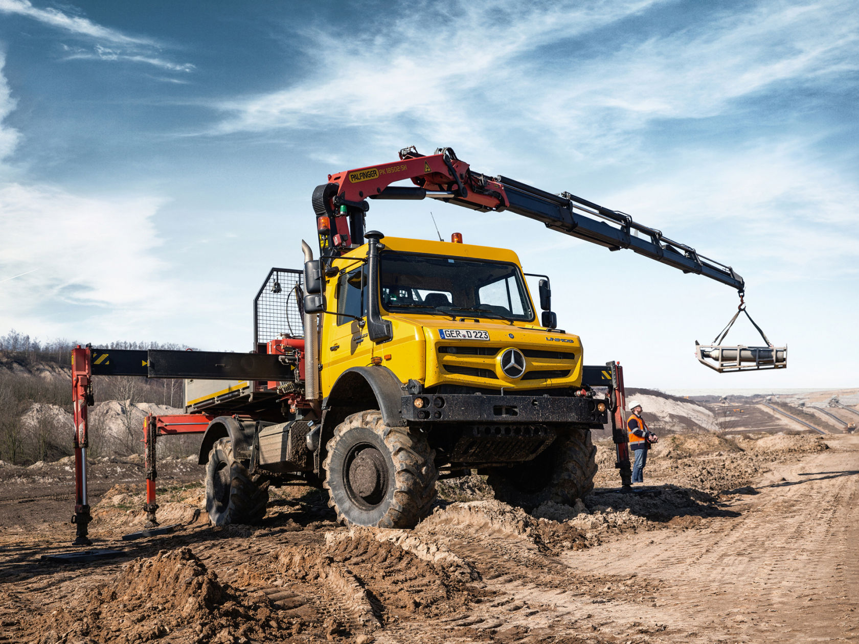 Un Unimog jaune avec grue soulève une charge dans une carrière ou un environnement similaire. Une personne en vêtements de travail se trouve à côté du véhicule.