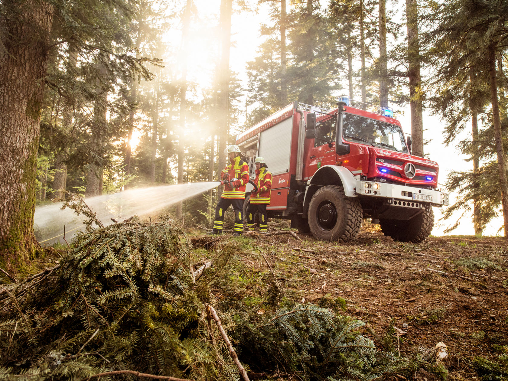 Un véhicule d'incendie rouge est stationné dans une forêt, tandis que les forces d'intervention pulvérisent de l'eau avec un flexible.