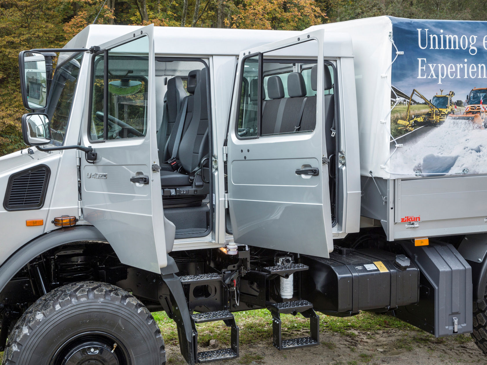 Ein silberner Unimog mit geöffneten Türen, die den Innenraum und die Sitze zeigen. Die Ladefläche des Lastwagens ist mit einer Plane bedeckt.