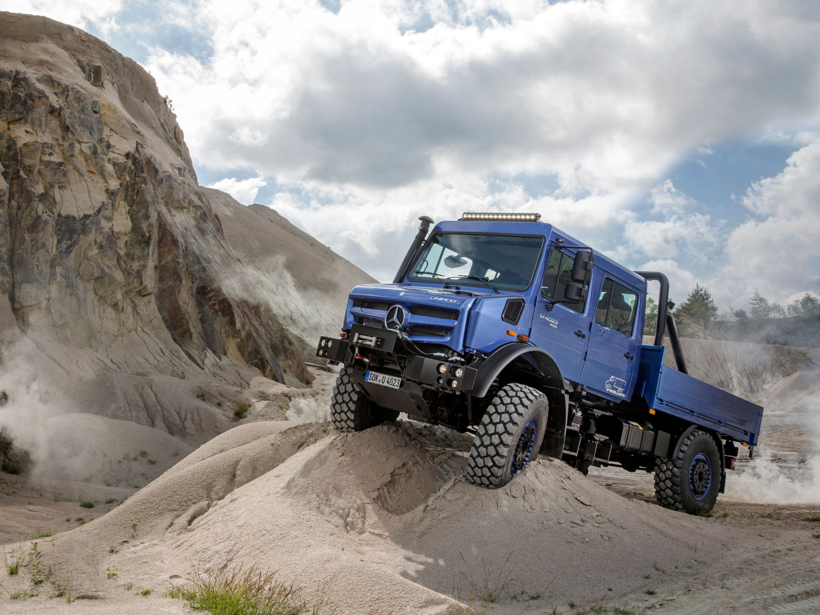 Een blauwe Unimog rijdt een steile zandduin op.