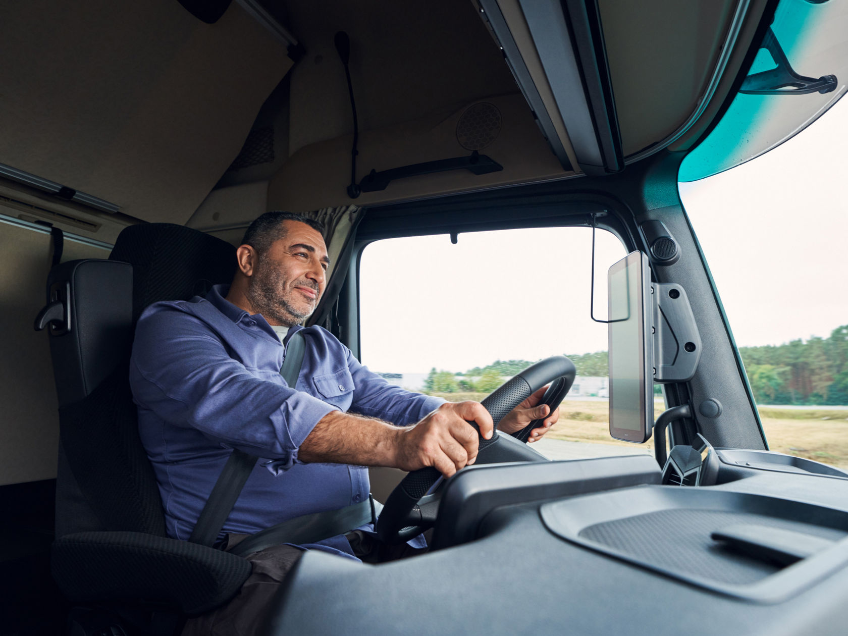 Een man met baard en blauw overhemd zit aan het stuur van een Actros L en glimlacht. Hij houdt het stuurwiel met beide handen vast terwijl hij op een weg rijdt die door bomen is omgeven.
