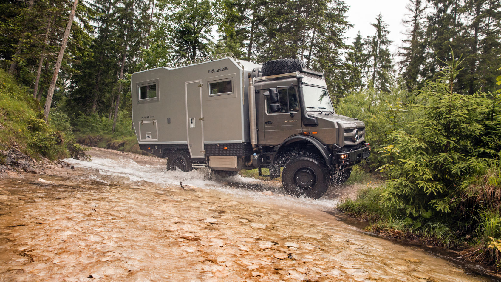 Das Bild zeigt einen grauen Unimog mit einem Wohnmobilaufbau, der einen Fluss durchquert. Das Fahrzeug ist seitlich zu sehen und spritzt Wasser auf, während es durch das flache Gewässer fährt. Der Hintergrund besteht aus einem dichten Wald mit hohen Bäumen und grüner Vegetation. Das Bild vermittelt ein Gefühl von Abenteuer und Offroad-Fähigkeit.