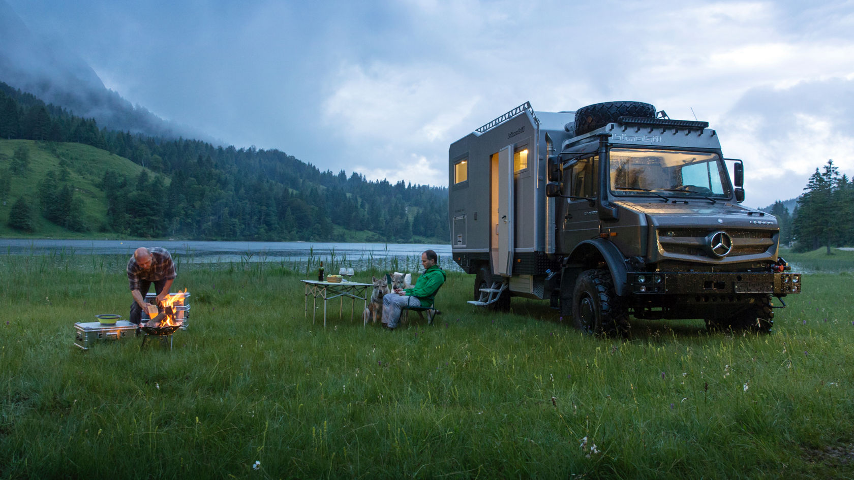Das Bild zeigt eine malerische Szene mit einem grauen Unimog Expeditionsmobil, das auf einer grünen Wiese am Ufer eines Sees geparkt ist. Zwei Männer sind zu sehen: Einer kümmert sich um ein Lagerfeuer, während der andere mit zwei Hunden an einem Tisch sitzt. Im Hintergrund erheben sich bewaldete Hügel, die in den bewölkten Himmel übergehen. Die Szene vermittelt ein Gefühl von Ruhe und Abenteuer in der Natur.