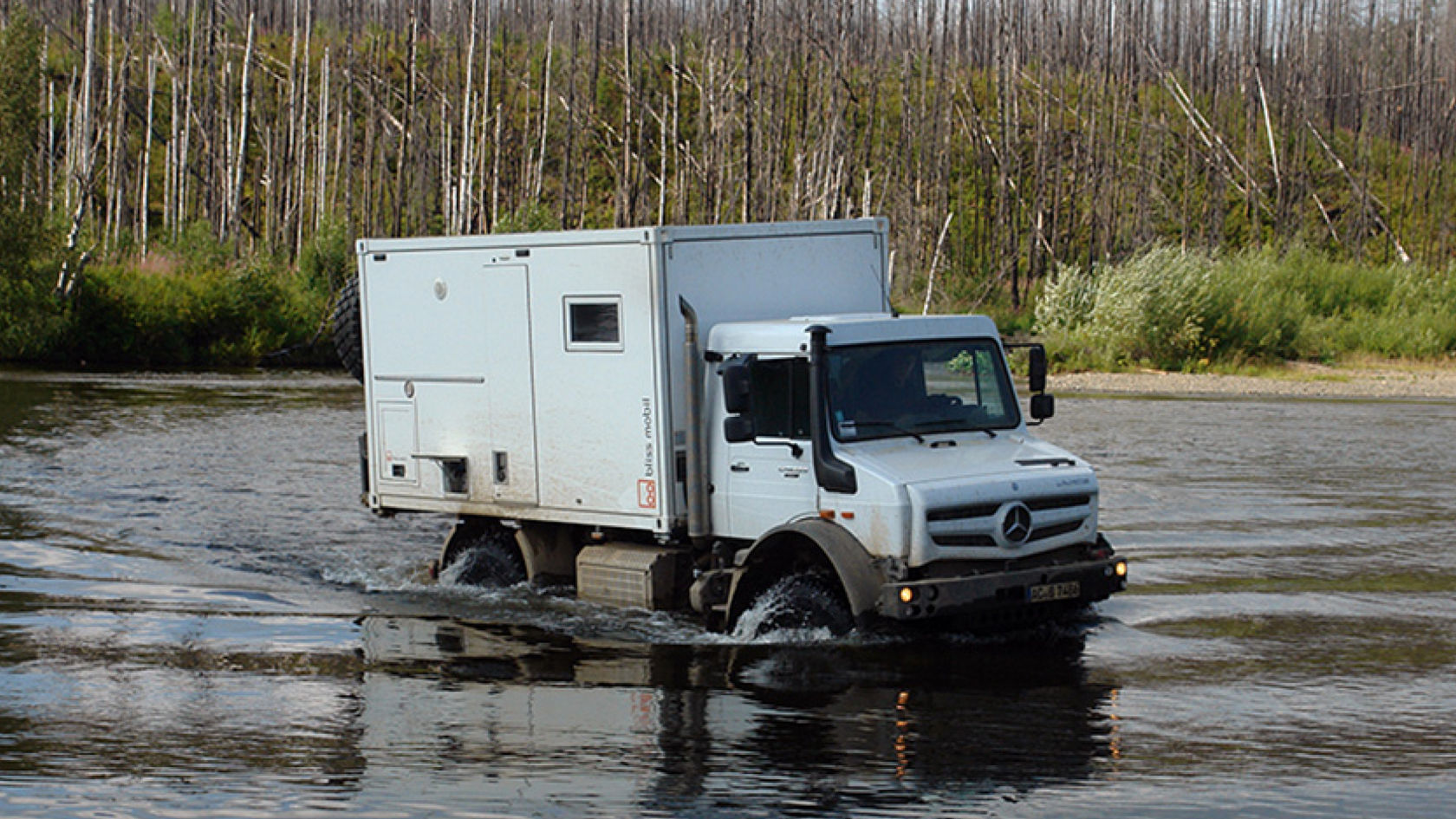 Das Bild zeigt einen weißen Unimog mit einem Aufbau, der wie ein Expeditionsmobil aussieht, der einen Fluss durchquert. Das Wasser spritzt um die Räder des Fahrzeugs, während es sich durch das flache Gewässer bewegt. Im Hintergrund sind Bäume und Büsche zu sehen, die den Fluss säumen.