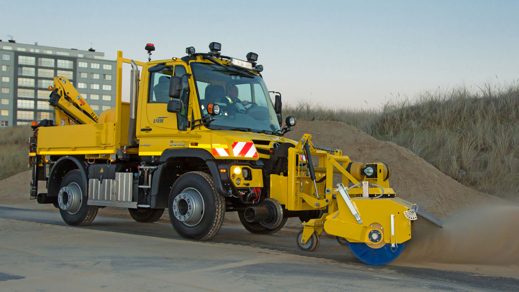Liiva eemaldamise erivarustusega kollane Unimog, kasutusel liivasel pinnal hoone ja luidete ees.