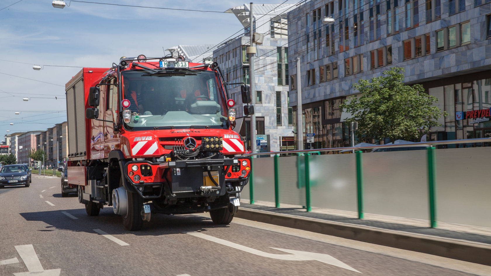 Rööbastee varustusega punane Unimog sõidab linnateel moodsate hoonete kõrval.