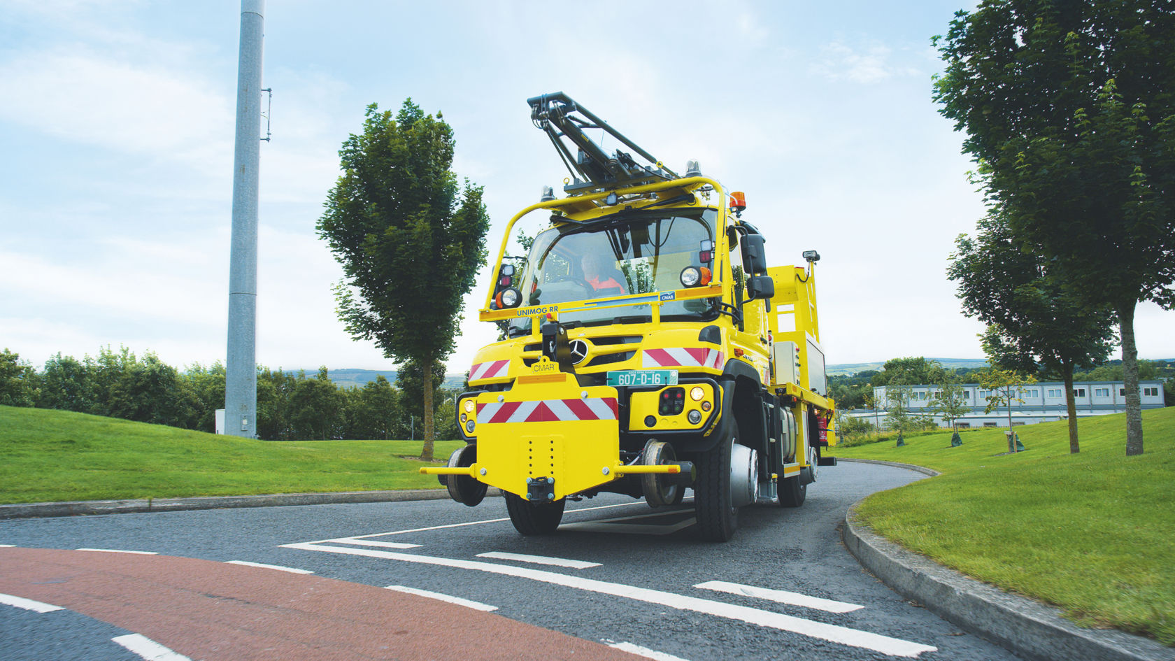 黄色 Unimog 带铁路设备，在带树木和草坪的绿色环境中行驶在道路上。