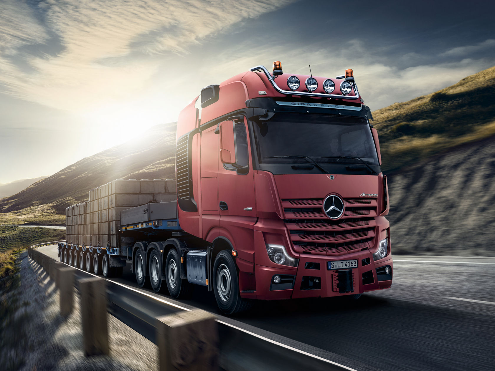 La photo montre un Actros rouge avec une remorque surbaissée chargée de grosses et lourdes pièces préfabriquées en béton. Le camion roule sur une route à travers un paysage vallonné. Le soleil brille et le ciel est nuageux. Le camion possède une cabine « GigaSpace » et est équipé de phares additionnels.