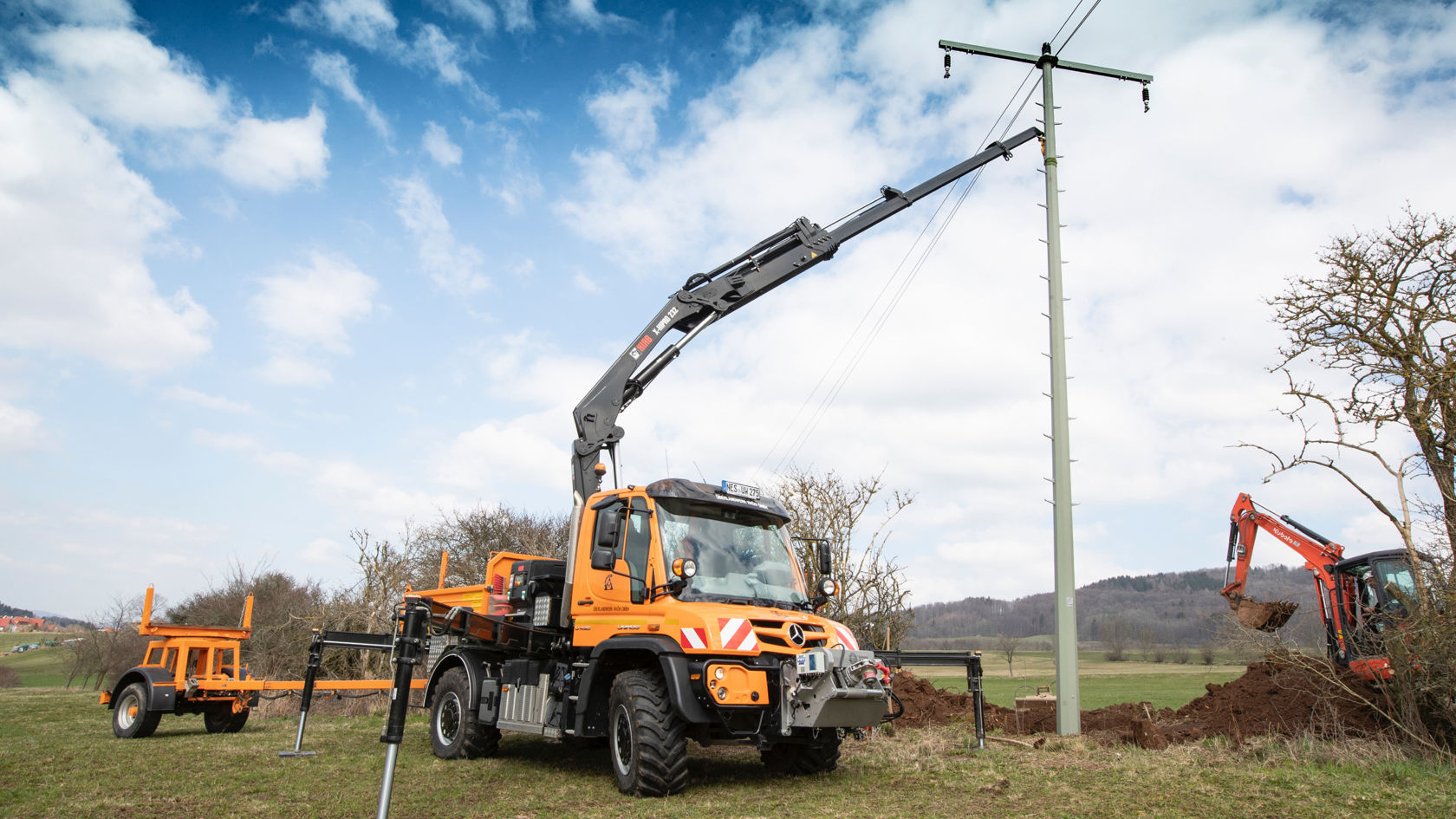 La photo montre un Unimog orange avec un bras de grue travaillant sur un poteau électrique. Le camion se trouve dans un champ herbeux et le bras de la grue est déployé pour atteindre le mât. Une remorque orange se trouve derrière le camion et une petite pelleteuse est visible en arrière-plan. Le ciel est bleu avec quelques nuages.