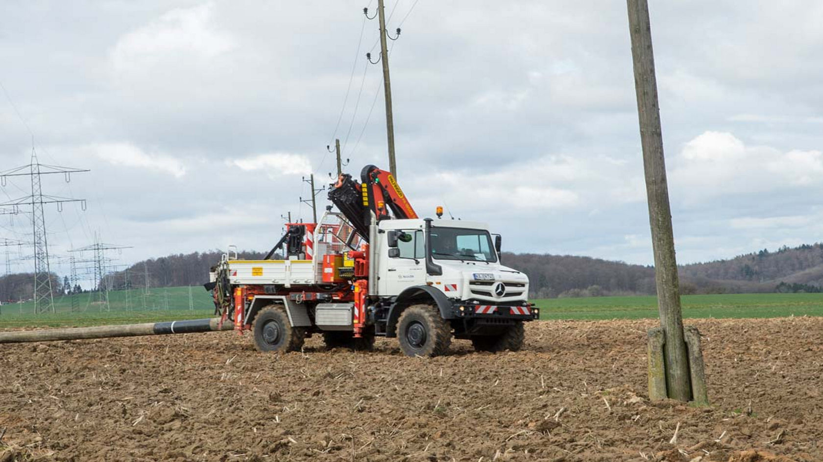 La photo montre un Unimog blanc équipé d'un bras de grue, stationné dans un champ. Le camion est équipé d'un bras de grue qui peut être utilisé pour soulever et déplacer des charges. En arrière-plan, on aperçoit des poteaux électriques et une zone boisée. Le ciel est nuageux.