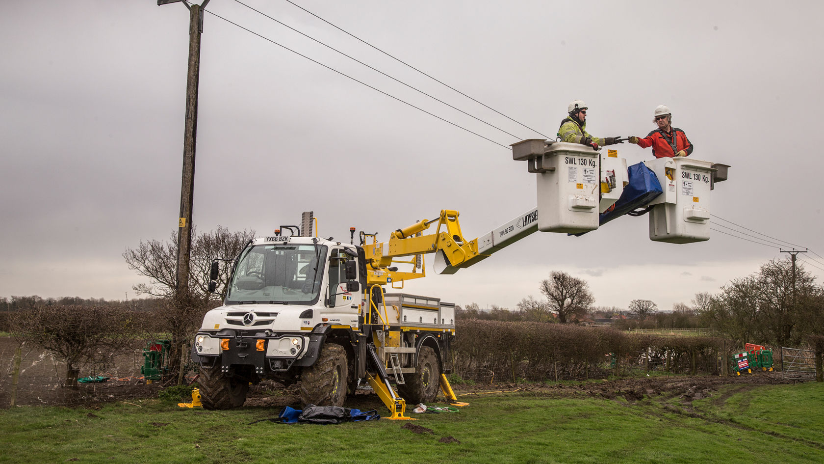 Sur la photo, on peut voir un Unimog blanc équipé d'un bras articulé jaune qui maintient deux ouvriers en suspension dans les airs. Les ouvriers portent des casques et semblent travailler sur des lignes électriques. Le camion est stationné dans un champ herbeux avec un poteau électrique à proximité. Le ciel est nuageux.