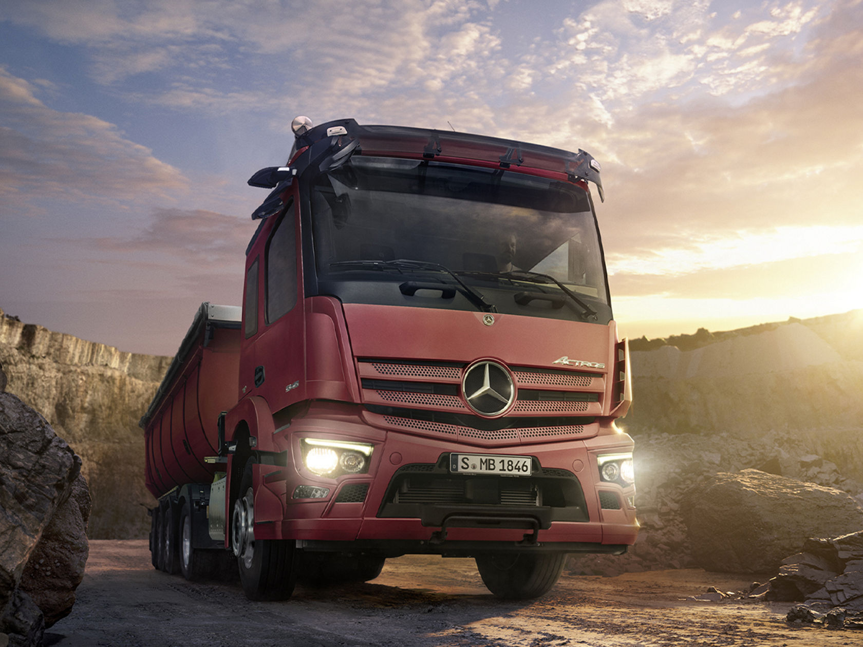 La photo montre un camion-benne Actros rouge stationné dans une carrière. Le camion est visible de face avec une vue latérale vers l'arrière. Ses phares sont allumés. En arrière-plan, on aperçoit des falaises et un ciel nuageux. Le soleil brille et projette une lumière chaude sur la scène.