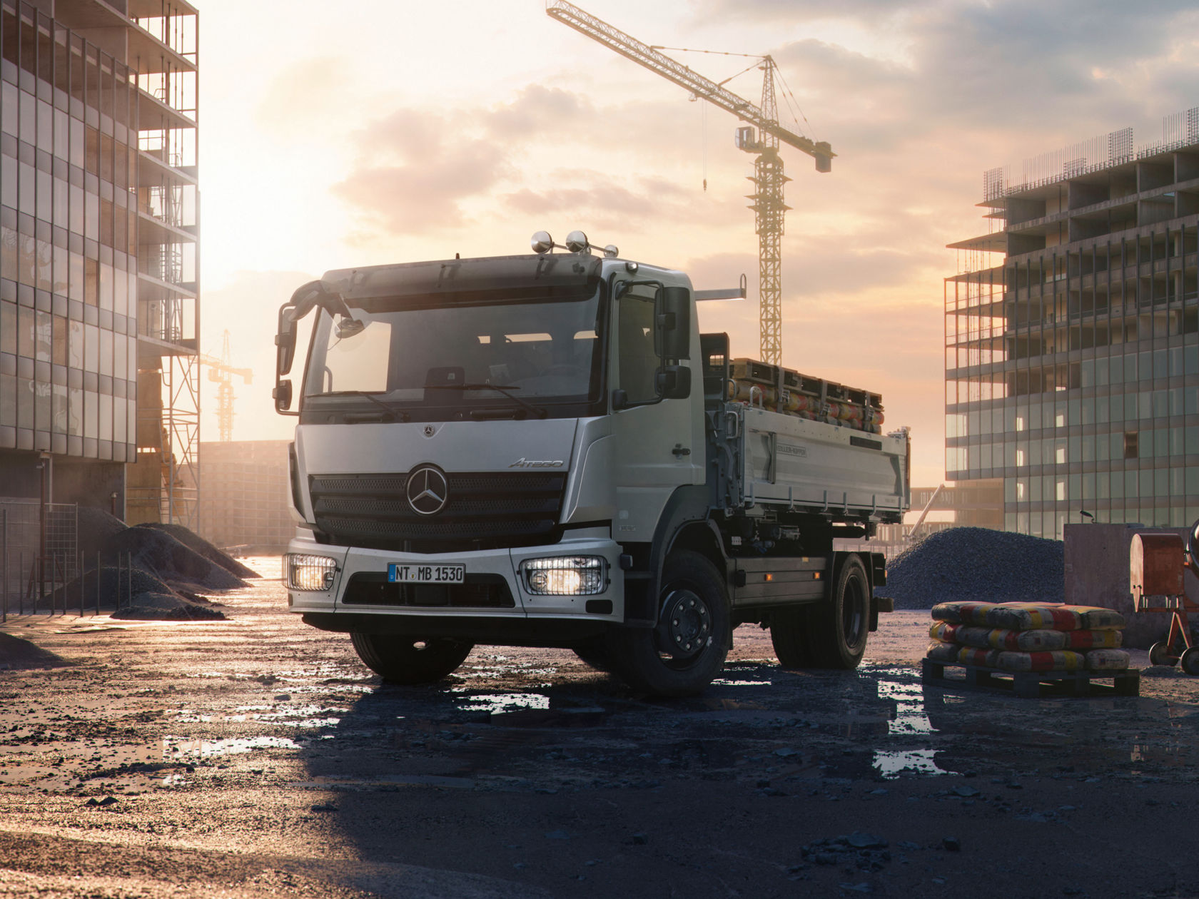 La photo montre un Atego blanc sur un chantier. Le camion est équipé d'une benne chargée de palettes. En arrière-plan, on aperçoit un bâtiment inachevé et une grue de chantier. Le ciel est clair et il semble être le soir. Des flaques et des matériaux de construction sont visibles au sol.