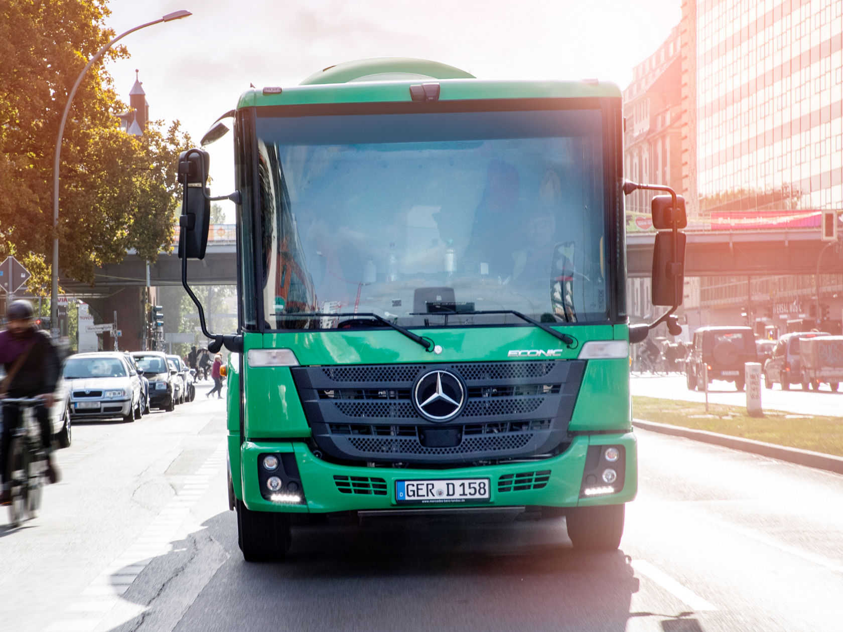 La photo montre un camion Econic vert circulant sur une route dans une ville. Le camion est visible de face. En arrière-plan, on aperçoit d'autres véhicules, des piétons et des bâtiments. Le soleil brille et la journée semble claire. Un cycliste roule sur le côté gauche de la photo.