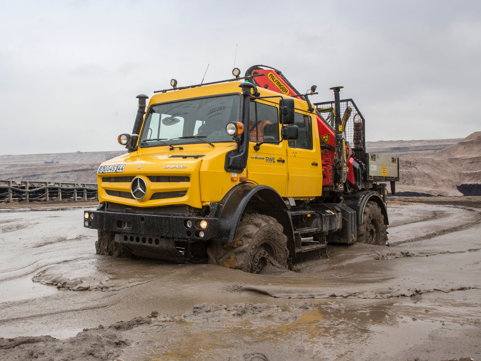 La photo montre un Unimog jaune avec un bras de grue rouge qui se trouve dans une zone boueuse. Les pneus du camion sont profondément enfoncés dans la boue. En arrière-plan, on aperçoit une mine à ciel ouvert et un ciel nuageux.