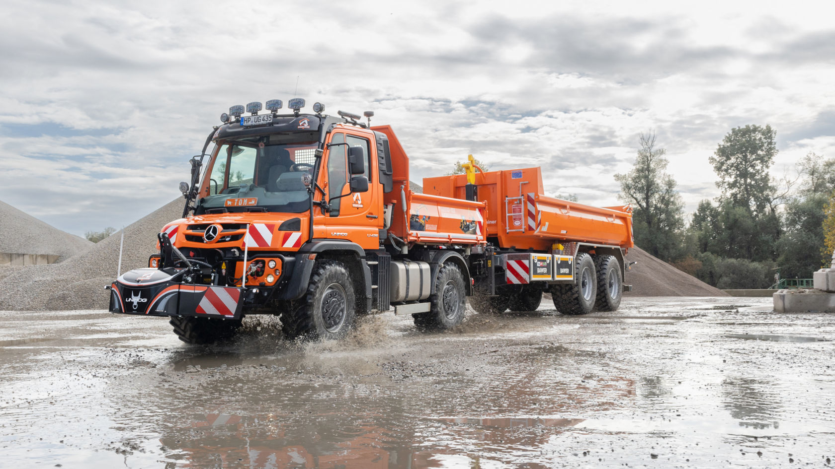 La photo montre un Unimog orange avec une remorque traversant une flaque d'eau. L'eau est projetée vers le haut et recouvre le camion. En arrière-plan, on aperçoit un tas de gravier et des arbres. Le ciel est nuageux.