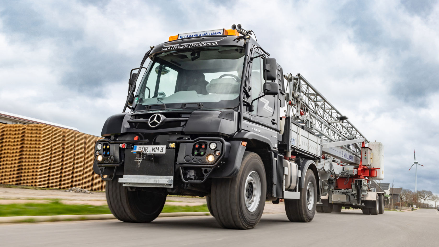 La photo montre un Unimog gris équipé d'une échelle télescopique sur une remorque, circulant sur une route. En arrière-plan, on peut voir une pile de palettes en bois, des maisons et une éolienne. Le ciel est nuageux.
