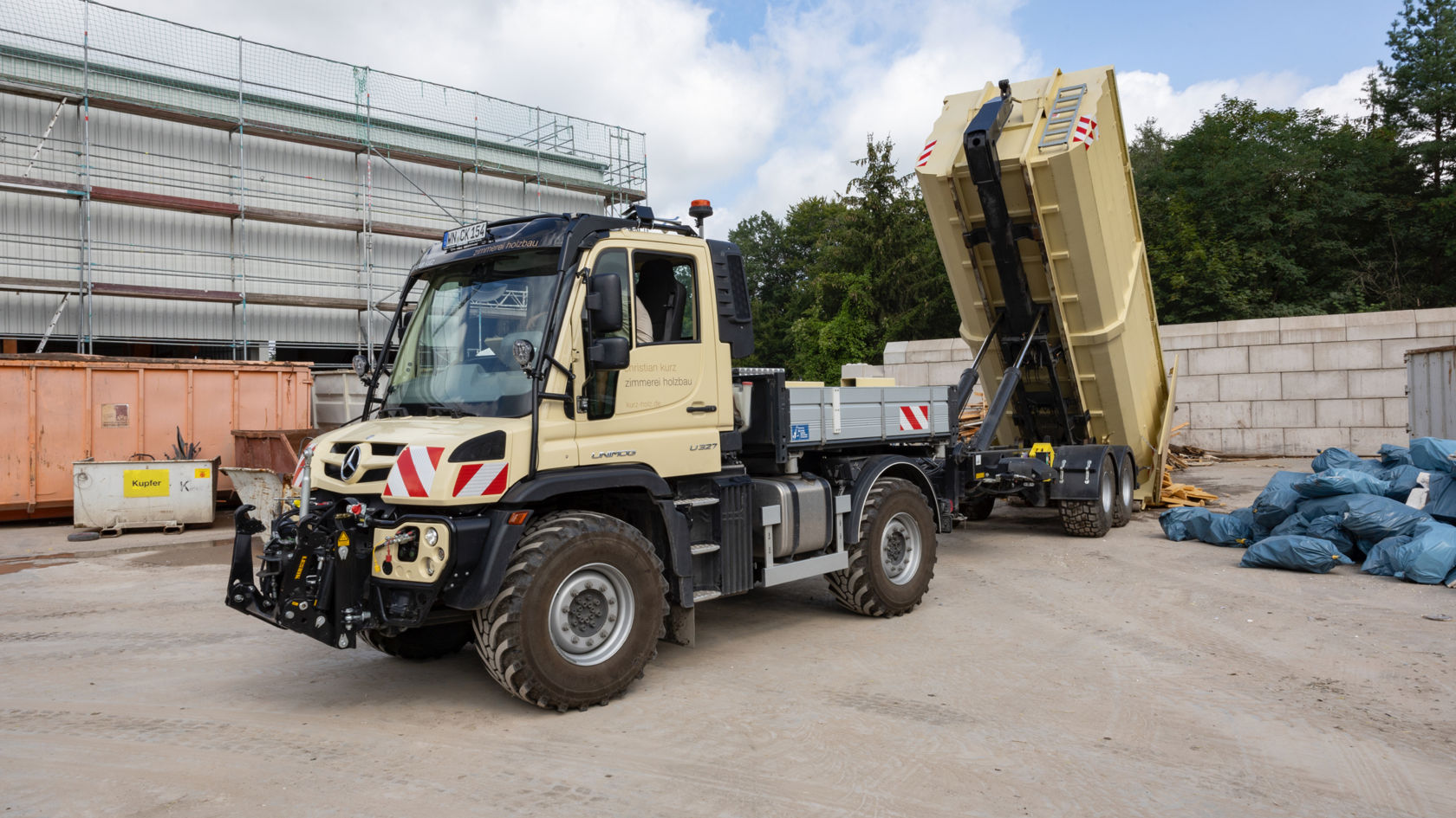 La photo montre un Unimog beige avec une remorque à benne basculante, stationné dans une cour. La remorque à benne basculante est relevée et décharge des déchets de bois. En arrière-plan, on peut voir un bâtiment avec un échafaudage, des conteneurs et des sacs poubelles bleus. Le ciel est nuageux.
