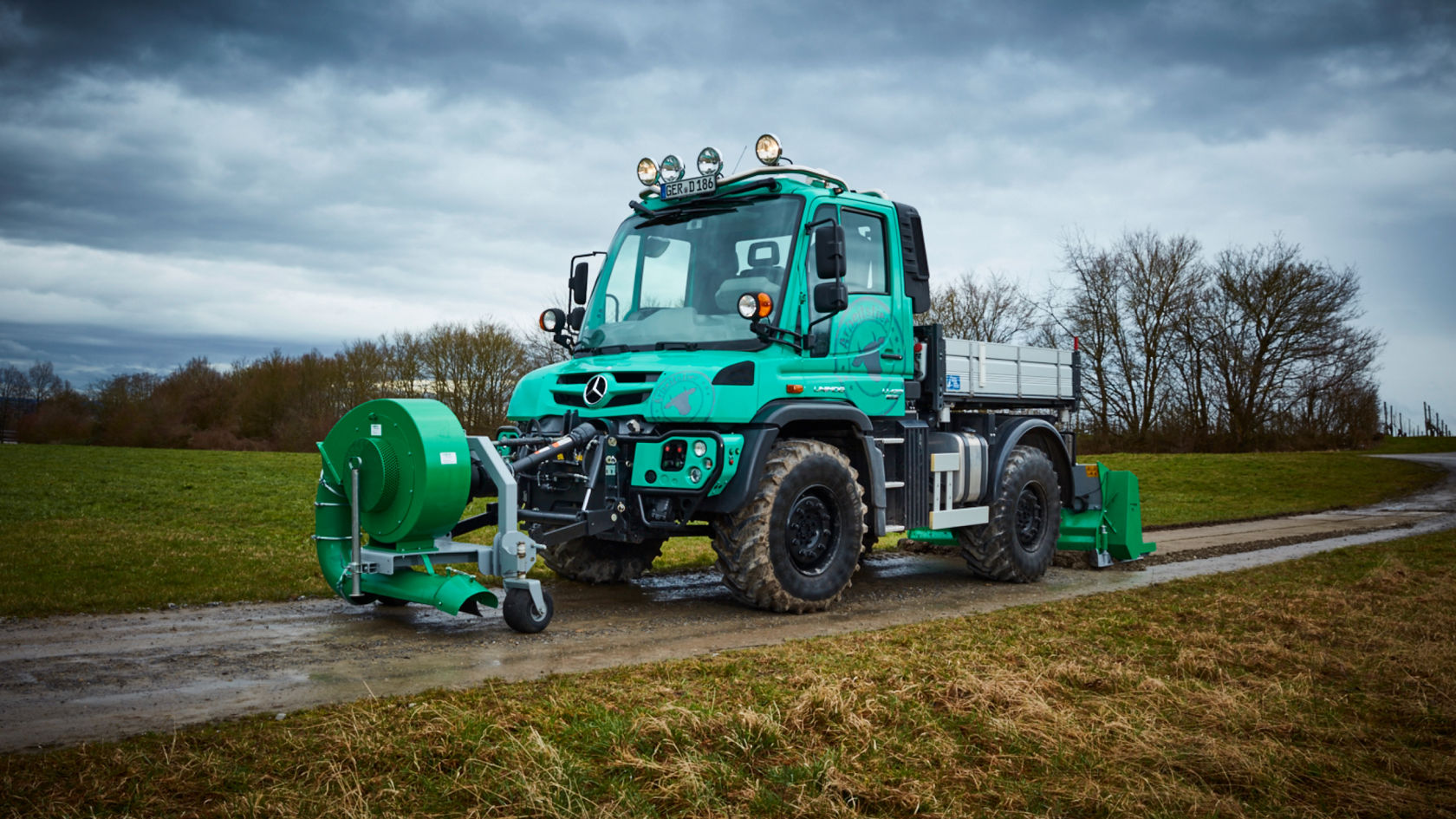 Un Unimog blanc roule sur un chemin boueux. Le camion projette de l'eau lorsqu'il traverse une flaque d'eau. En arrière-plan, on peut voir des champs et des arbres.