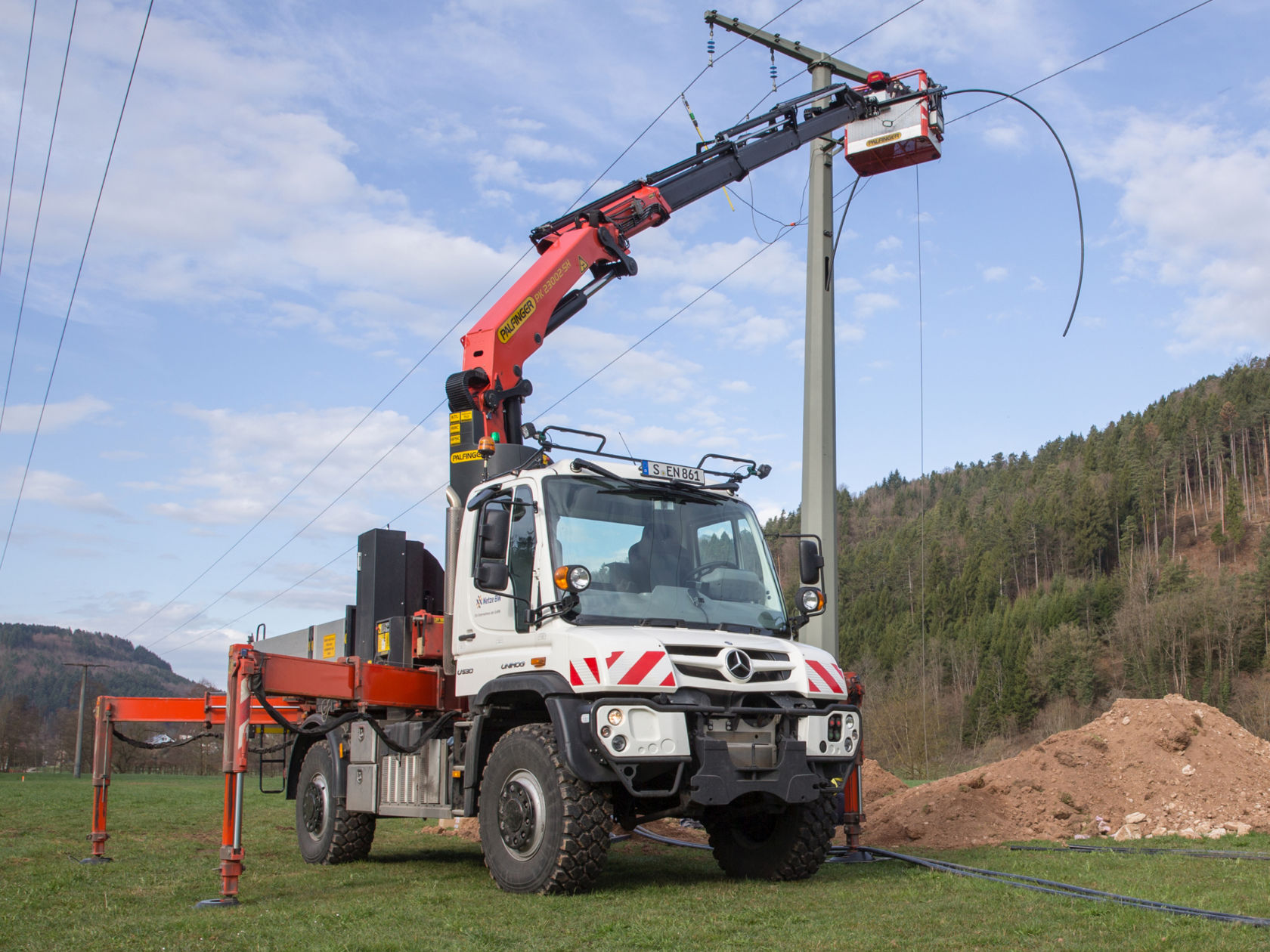 La photo montre un Unimog blanc avec un bras de grue rouge travaillant sur un poteau électrique. Le bras de grue est équipé d'une plate-forme élévatrice dans laquelle se trouve une personne. Le camion se trouve sur un champ herbeux, avec une forêt et un ciel bleu en arrière-plan.