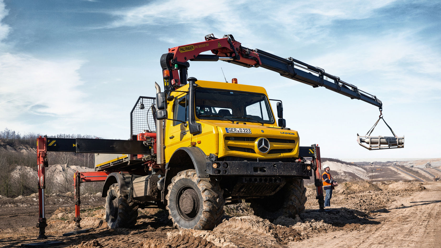La photo montre un Unimog jaune avec un bras de grue rouge, stationné sur un chantier. Le bras de grue soulève une charge. Un ouvrier du bâtiment portant un casque et un gilet de sécurité se tient près du camion. En arrière-plan, on aperçoit une colline et un ciel bleu.