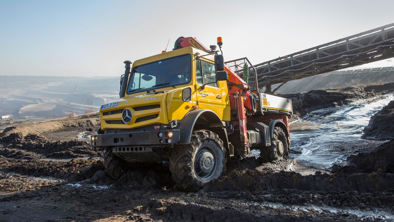 Un Unimog jaune sur un terrain boueux. Le camion est équipé d'un bras de grue rouge et semble être conçu pour des travaux lourds. Un pont ou un convoyeur est visible en arrière-plan.