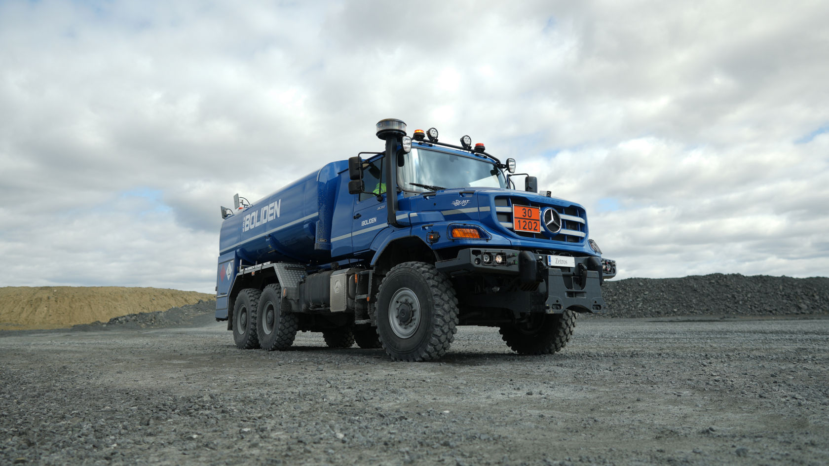 Un camion-citerne Zetros bleu est stationné sur un terrain gravelé. Le camion est équipé de grands pneus et est conçu pour une utilisation en tout-terrain. En arrière-plan, on peut voir des collines composées de terre et de pierres. Le ciel est nuageux.