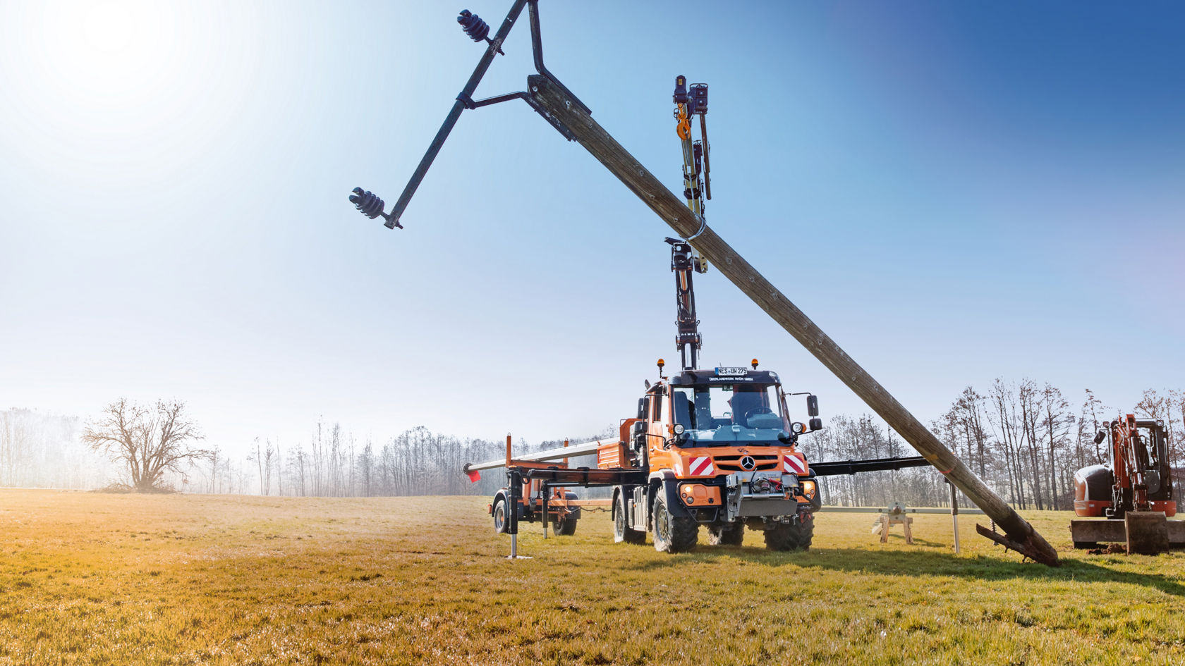 La photo montre un Unimog orange équipé d'un bras de grue qui soutient un poteau électrique. Le camion se trouve dans un champ herbeux et le poteau électrique est incliné sur le sol. Une remorque est attelée au camion. En arrière-plan, on peut apercevoir des arbres et un ciel bleu. Une petite pelleteuse se trouve à droite du poteau électrique.
