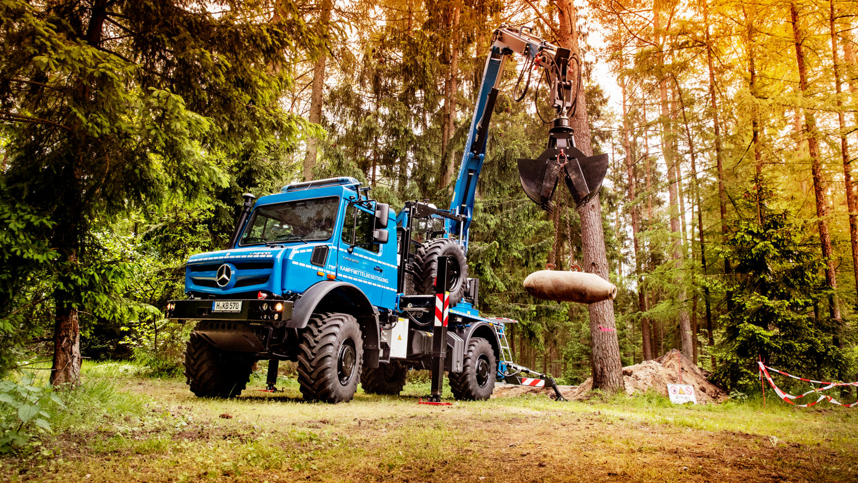 La photo montre un Unimog bleu avec un bras de grue, stationné en forêt. Le bras de grue tient une bombe. En arrière-plan, on aperçoit des arbres et une barrière.