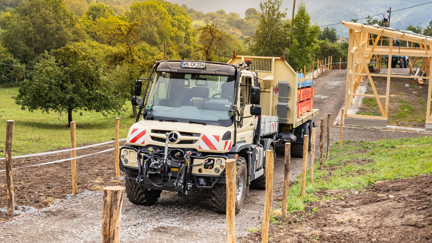 La photo montre un Unimog beige avec une remorque circulant sur un chemin de campagne. Le camion est pourvu de bandes rouges et blanches. En arrière-plan, on peut voir des arbres, une construction en bois et un ciel nuageux.