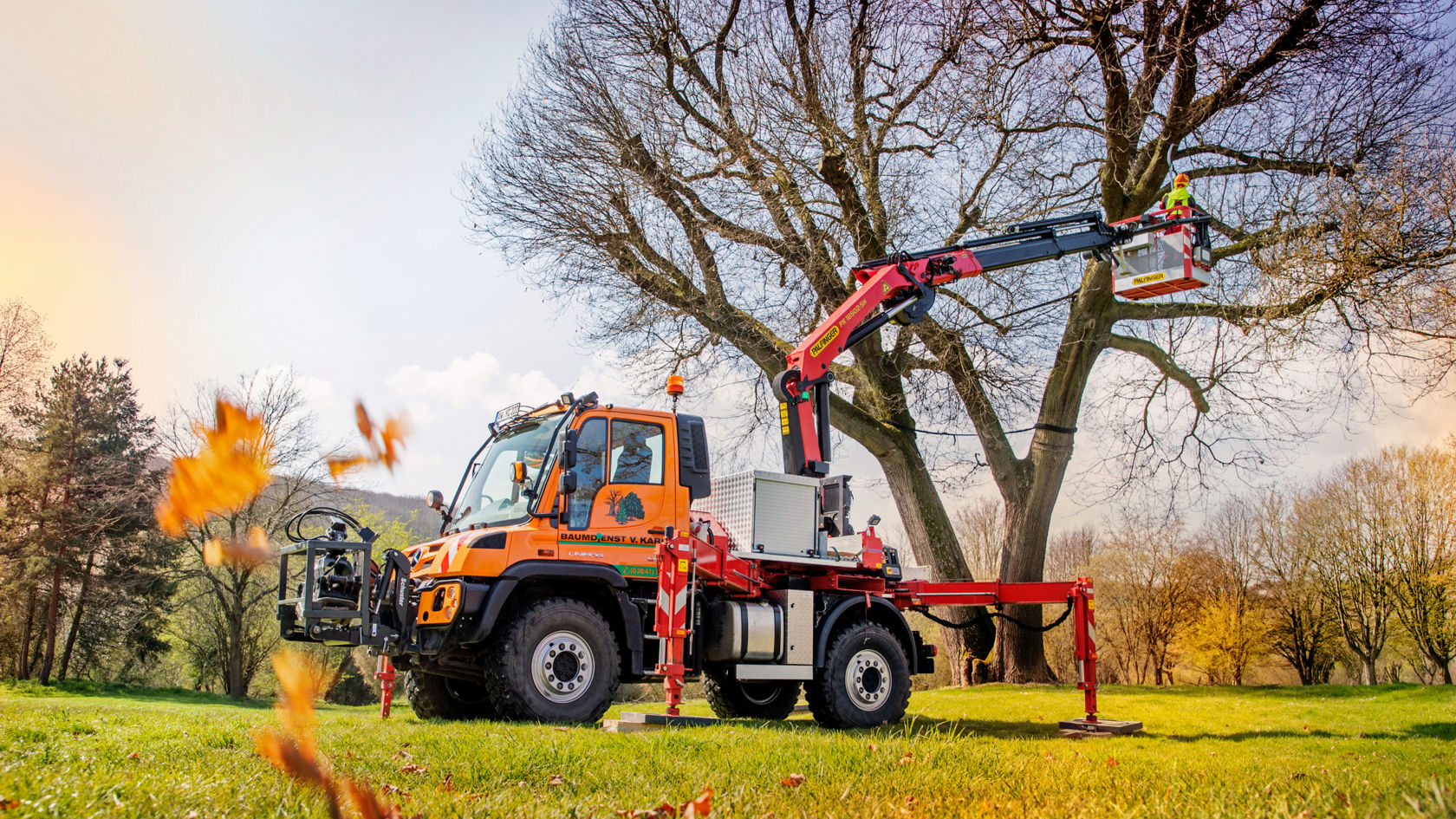 La photo montre un Unimog orange équipé d'une grue Palfinger. Il est utilisé pour des travaux d'entretien des arbres. Un ouvrier se trouve dans une nacelle à l'extrémité de la grue et travaille sur un grand arbre. Le camion est stationné dans un champ verdoyant, et des feuilles tombent au premier plan. Le soleil brille et le ciel est bleu.