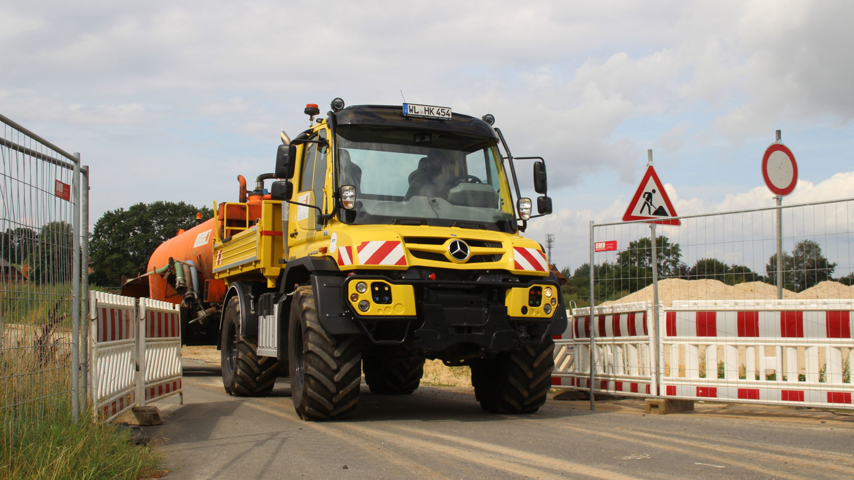 La photo montre un Unimog jaune avec une superstructure citerne orange. Le camion roule sur une route délimitée par des clôtures et des panneaux de signalisation. En arrière-plan, on peut apercevoir des arbres et un ciel nuageux.