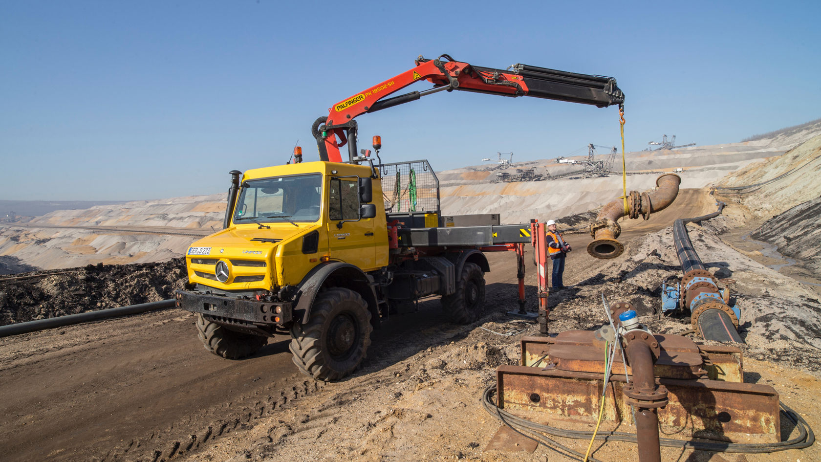 La photo montre un Unimog jaune avec une grue Palfinger dans un environnement minier. La grue soulève un grand tube alors qu'un ouvrier en vêtements de sécurité se trouve à côté. En arrière-plan, un vaste paysage composé de monticules de terre et d'équipements miniers s'étend sous un ciel bleu clair. D'autres tubes et appareils sont visibles sur le sol, ce qui suggère un chantier ou une zone de maintenance.
