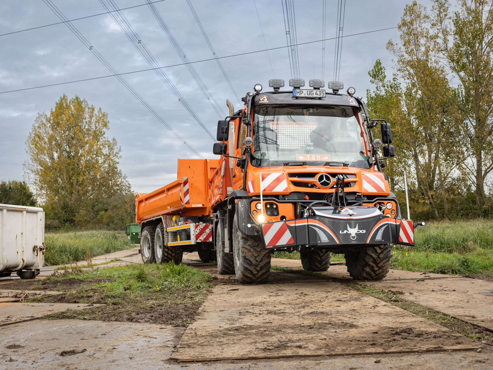 La photo montre un Unimog orange roulant sur une surface boueuse. Le camion est équipé d'un plateau basculant et d'un équipement frontal. Quatre phares additionnels se trouvent sur le toit du camion. En arrière-plan, on aperçoit des lignes électriques et des arbres. Le ciel est nuageux.