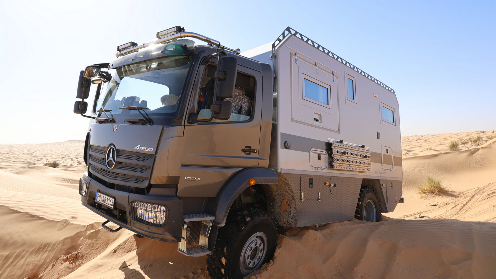Das Bild zeigt einen grauen Atego mit einem Aufbau, der wie ein Expeditionsmobil aussieht, in einer Wüstenlandschaft. Das Fahrzeug steht teilweise im Sand, und im Hintergrund sind Sanddünen unter einem hellen, sonnigen Himmel zu sehen. Der Atego ist mit zusätzlichen Scheinwerfern auf dem Dach ausgestattet.