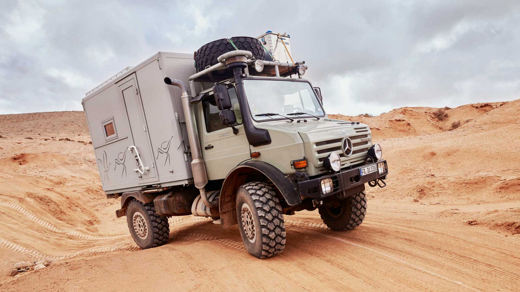 Das Bild zeigt einen hellgrauen Unimog mit einem Wohnmobilaufbau, der durch eine sandige Wüstenlandschaft fährt. Das Fahrzeug ist mit einem Reserverad auf dem Dach und zusätzlichen Scheinwerfern ausgestattet. Im Hintergrund sind sanfte Hügel und ein bewölkter Himmel zu sehen.