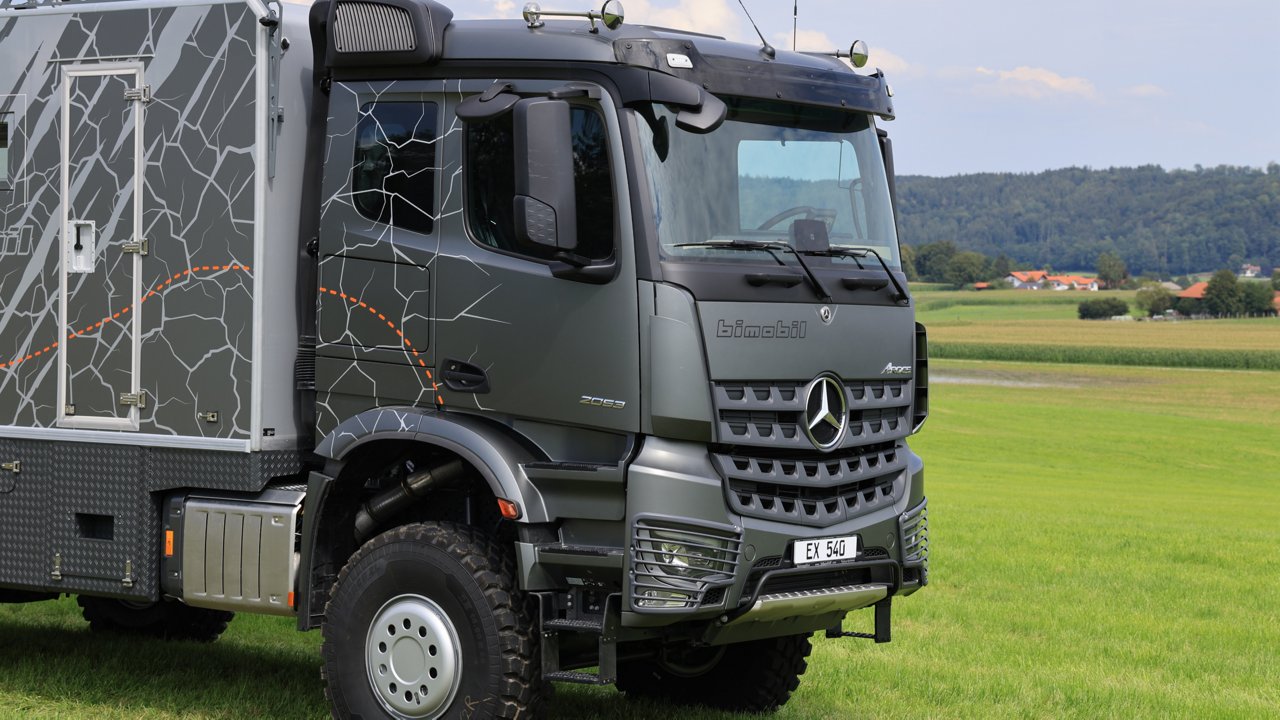 Das Bild zeigt einen grauen Arocs mit einem Expeditionsaufbau, der auf einer grünen Wiese steht. Das Fahrzeug hat ein markantes Design mit einem Muster, das an Risse oder Kartenlinien erinnert. Im Hintergrund sind Felder, Häuser und bewaldete Hügel zu sehen.